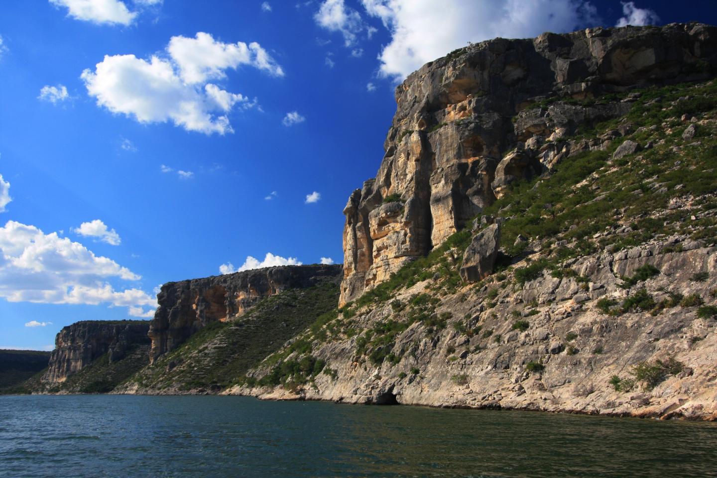 Steep rocky cliffs beside calm water under a blue sky with clouds.