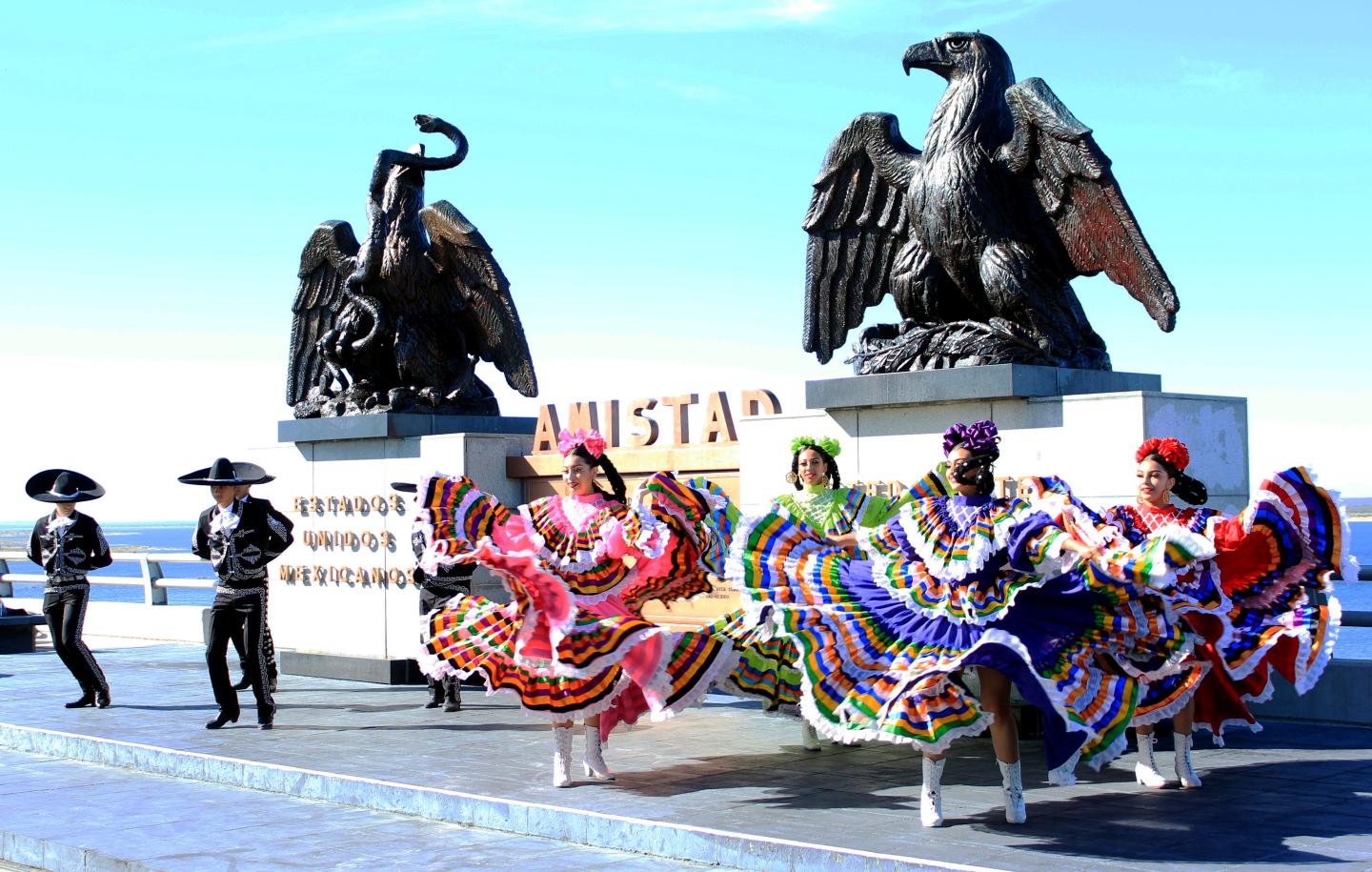 Dancers in colorful dresses perform near eagle statues under a clear sky.