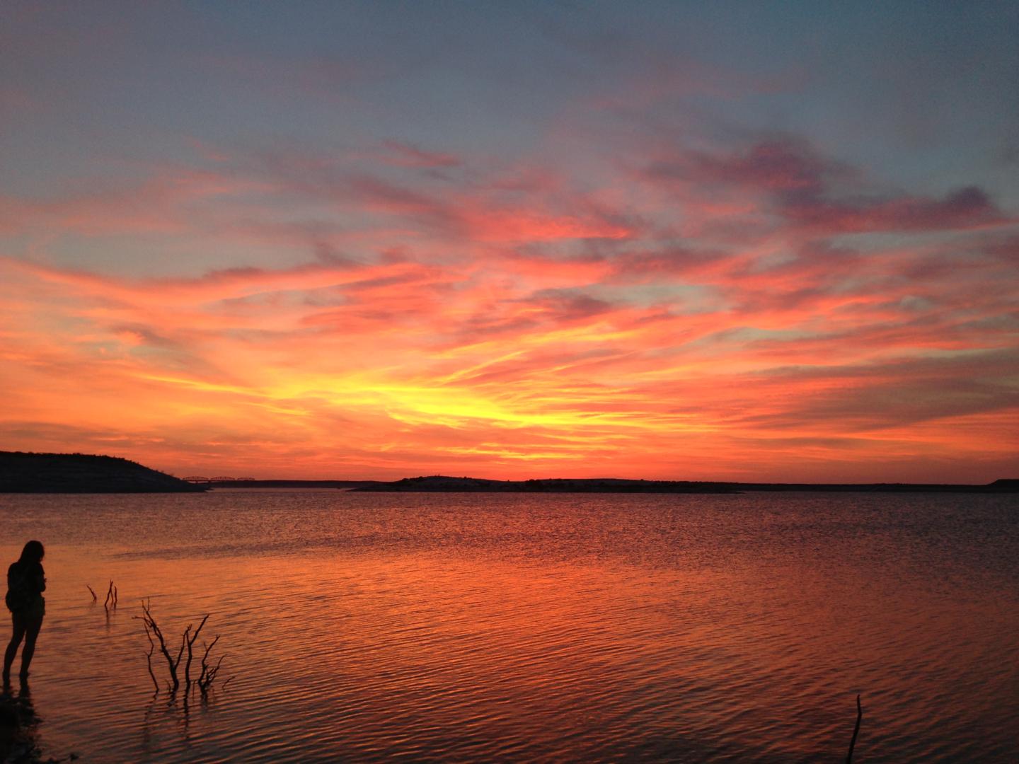 Sunset over a calm lake with a silhouetted person standing on the shore.