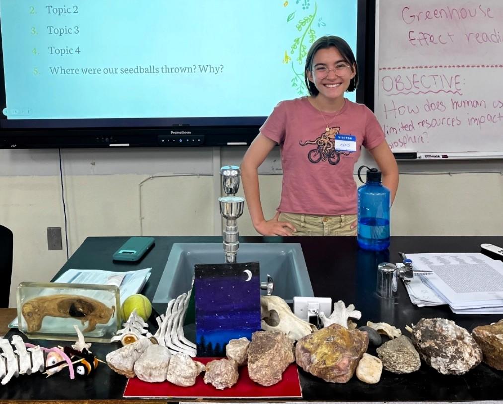 Smiling person stands behind a classroom table with rocks and fossils.