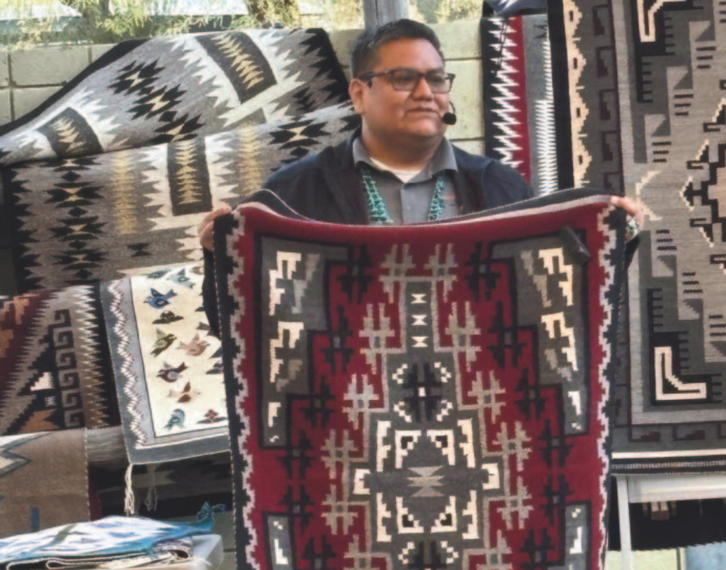 Hubbell Trading Post Trader, Wallace James Jr. showcasing a colorful Navajo rug with geometric designs in burgundy, gray, black, and white.
