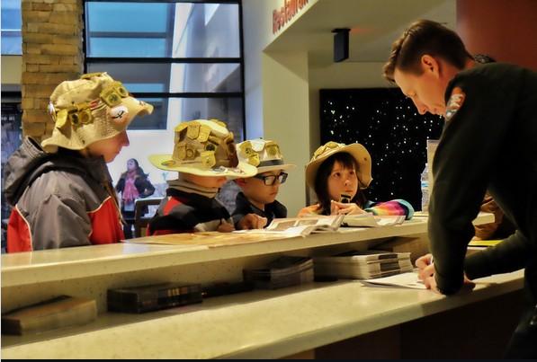 Group of kids with hats listening to a guide at a counter indoors.