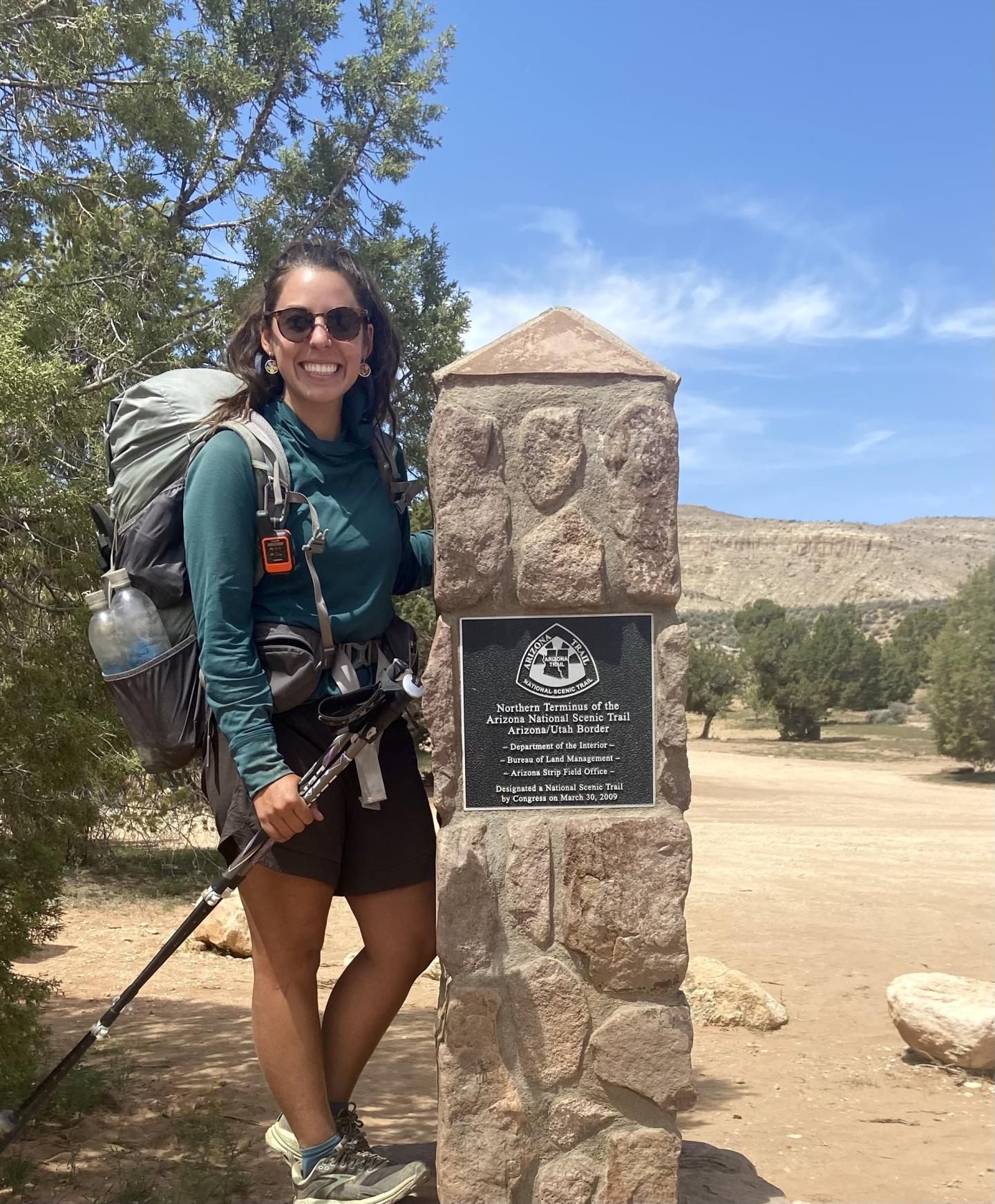Hiker smiling beside a stone marker in a sunny, open landscape.