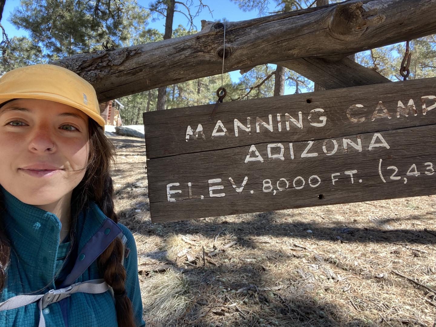 Smiling person by Manning Camp sign in a forest.