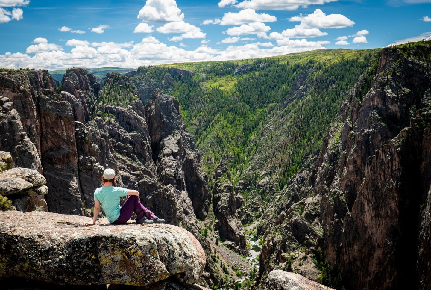 Person sitting on rock cliff, overlooking a lush, green canyon under a bright blue sky.