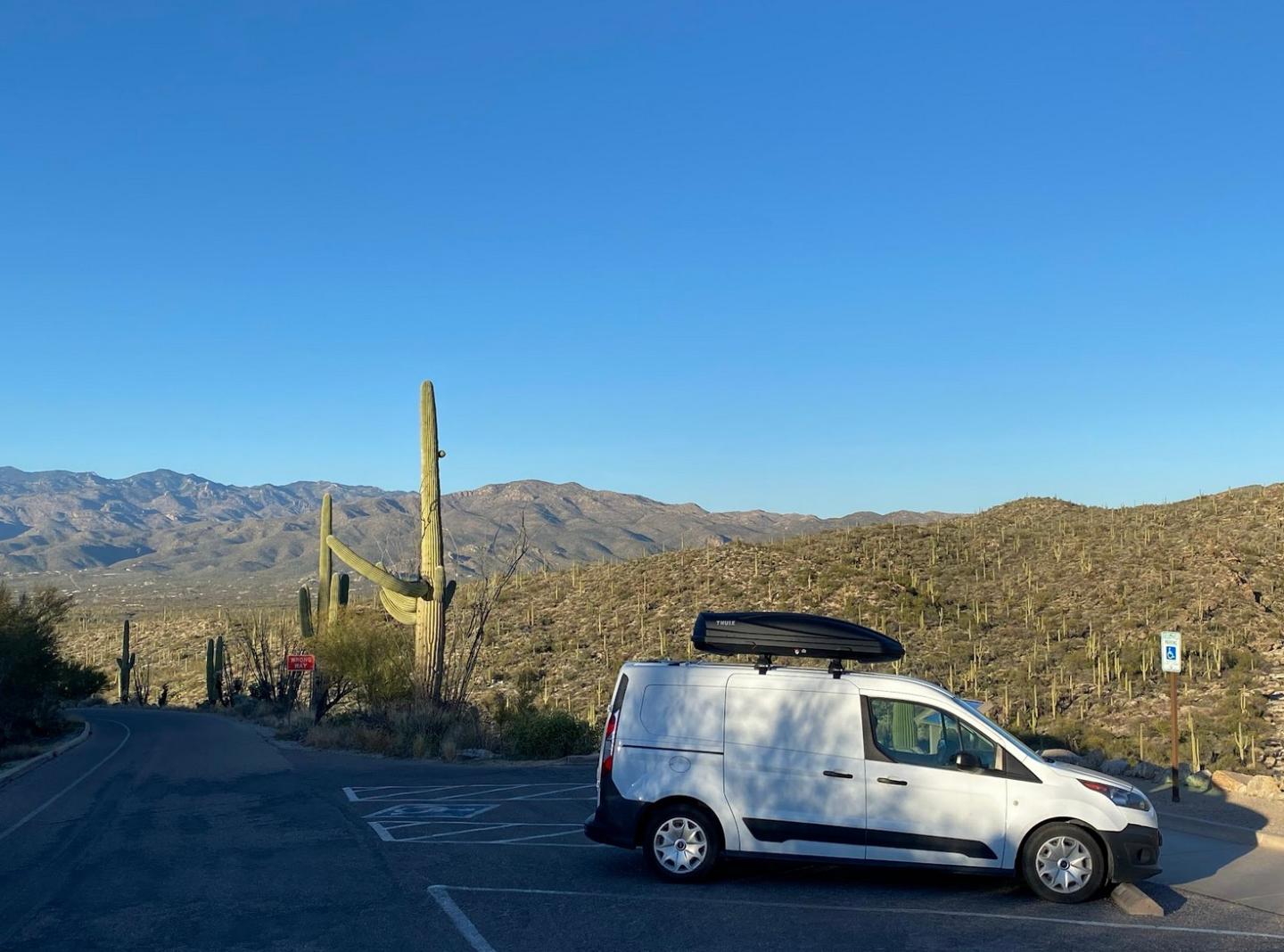 White van parked on a desert road, mountains and cacti in the background under clear blue sky.