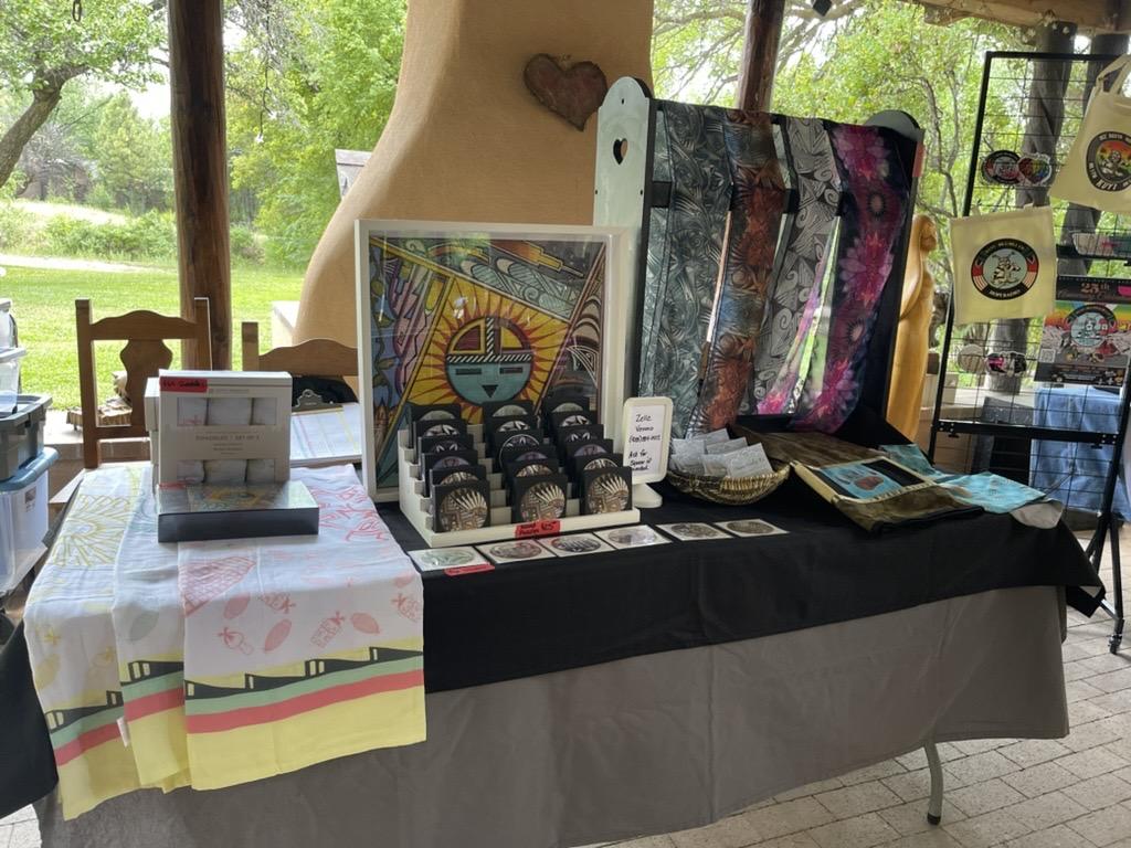 Craft fair table with colorful textiles and decorated boxes under a wooden shelter.