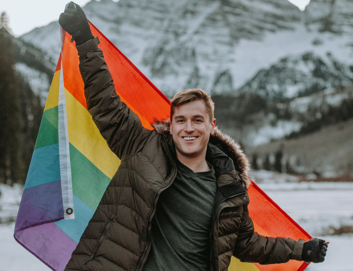 Smiling young, male person, film maker Mikah Meyers, holding a pride flag in a snowy mountain landscape.