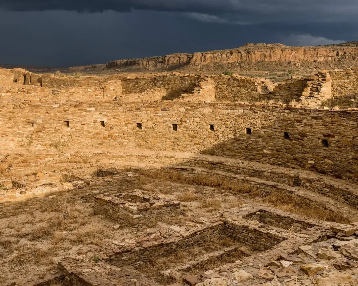 Ancient stone structure under a stormy sky, surrounded by rocky hills.