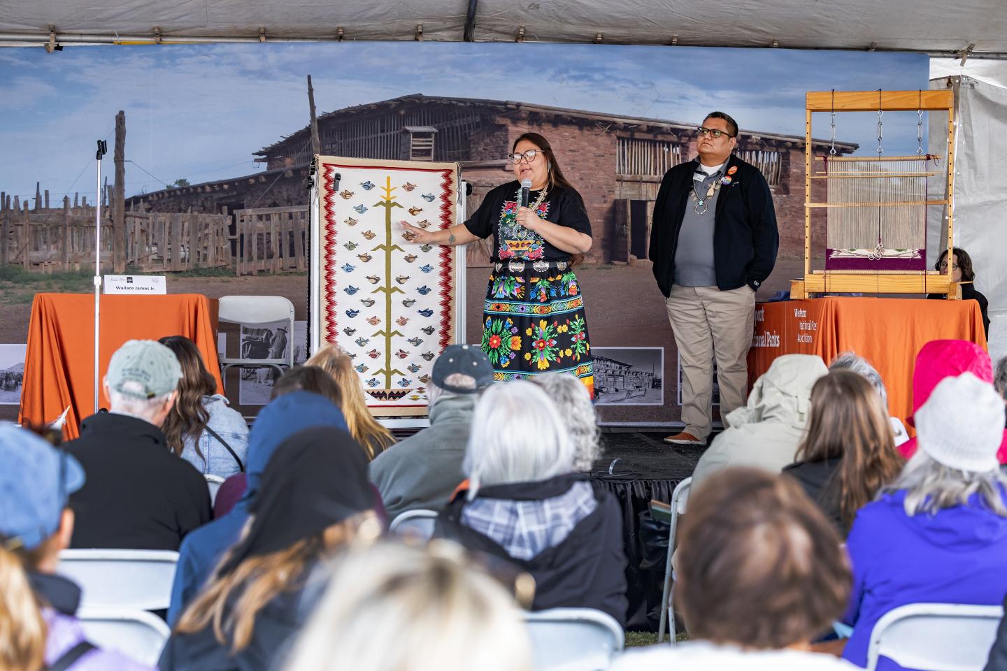 Two presenters in colorful attire on stage, addressing an outdoor audience.