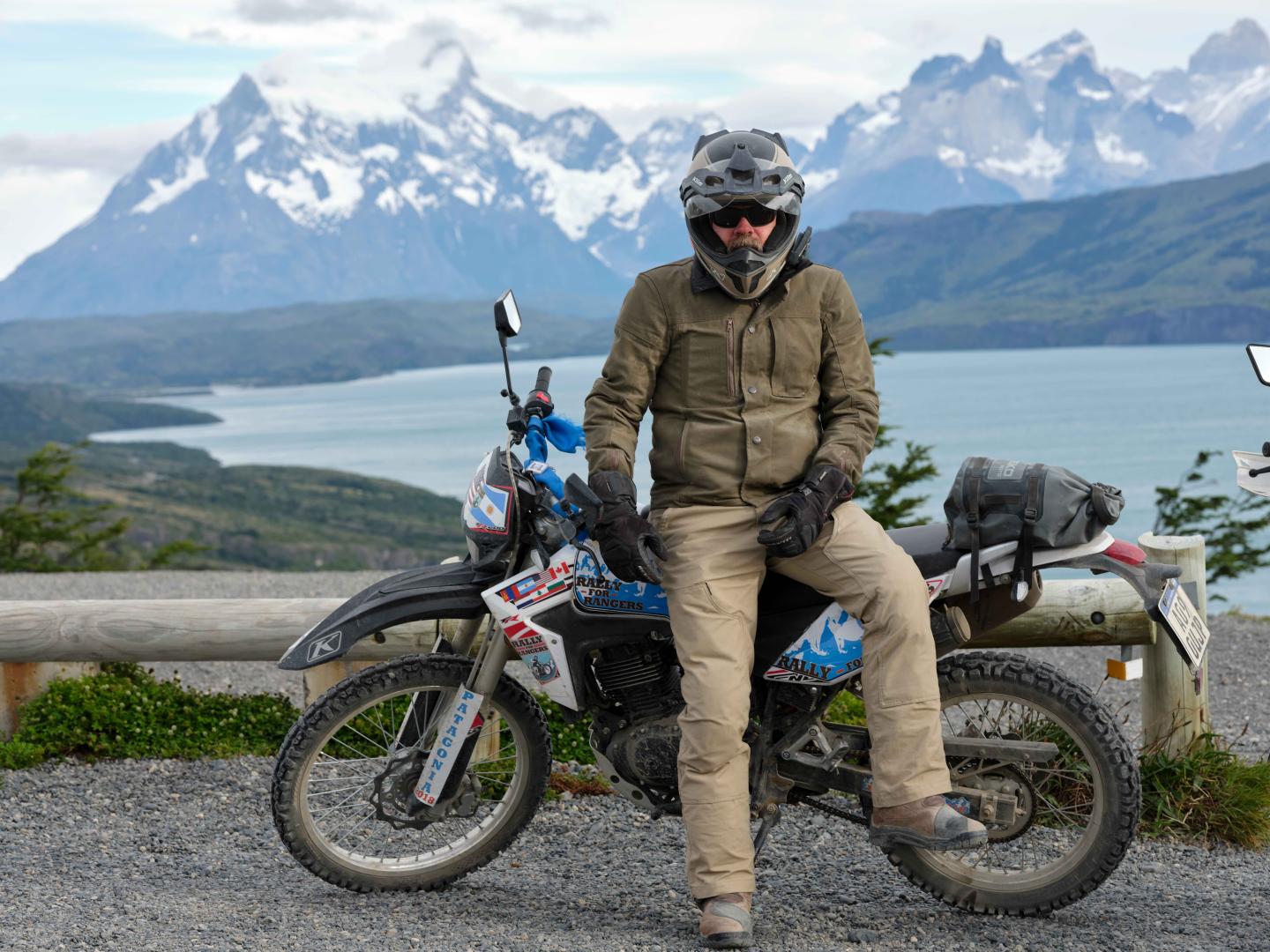 Man in riding gear sits on a motorcycle by a scenic lake and mountain backdrop.