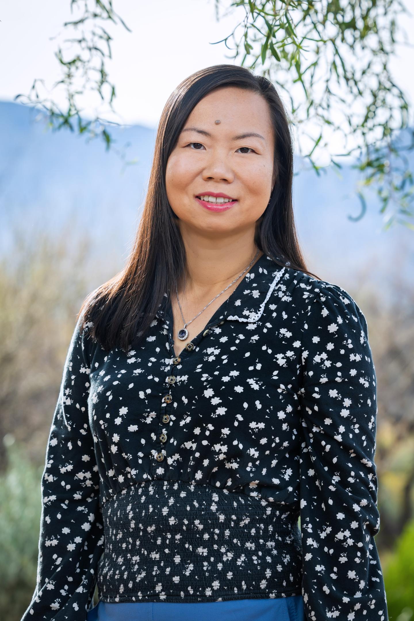 Smiling woman in a patterned top outdoors with trees and mountains in the background.