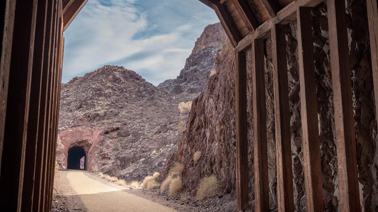 View from a wooden tunnel toward rocky, arid landscape under blue sky.