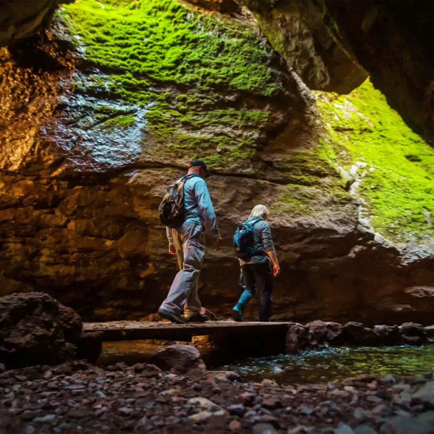 Two hikers walking through a moss-covered cave with sunlight streaming in.