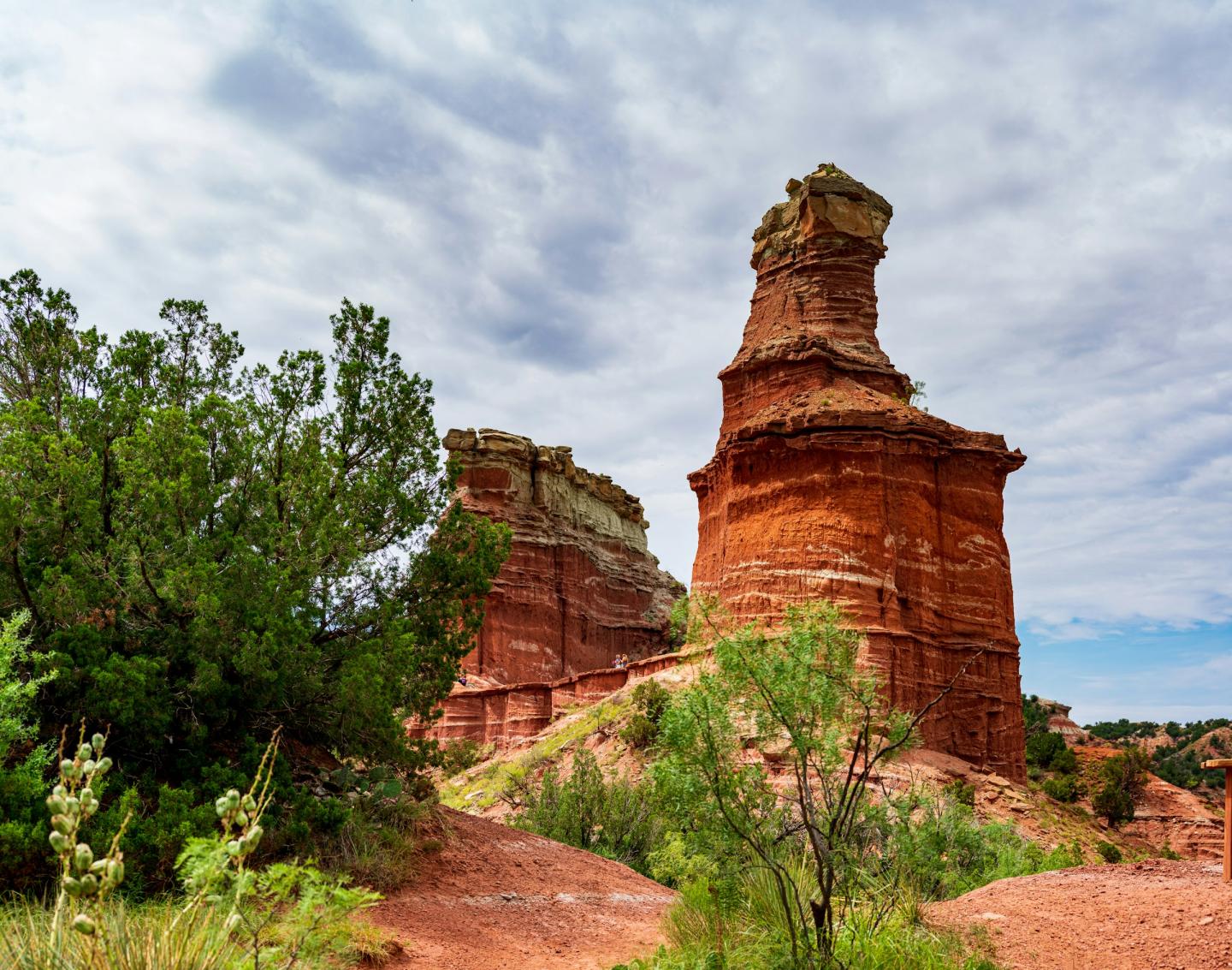 Red rock formations under a cloudy sky, with green shrubs in the foreground.