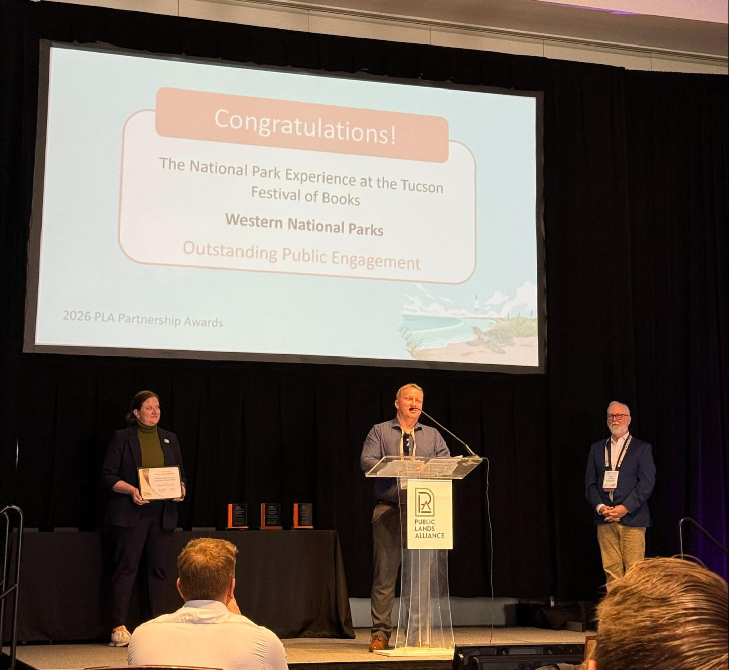 Three people on stage at an award ceremony, one speaking at podium.