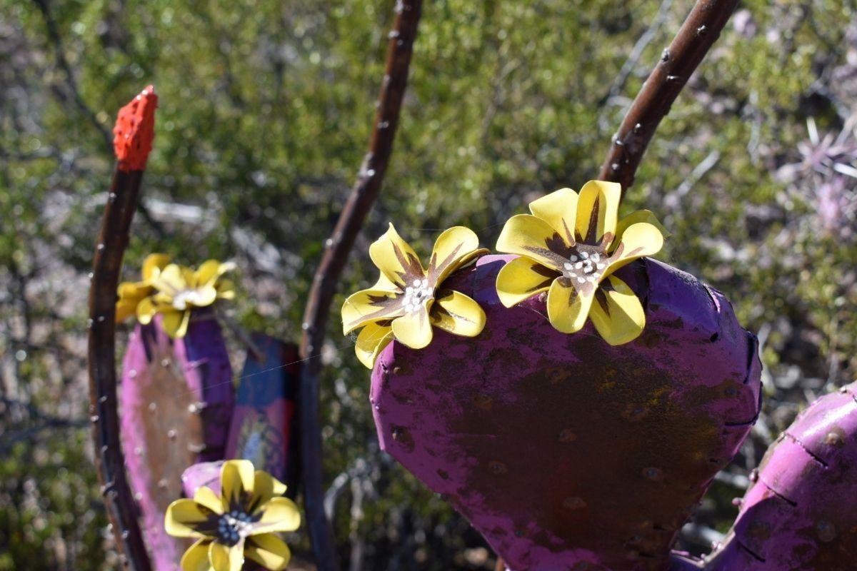 Purple cactus sculpture with yellow flowers, set in a desert landscape.
