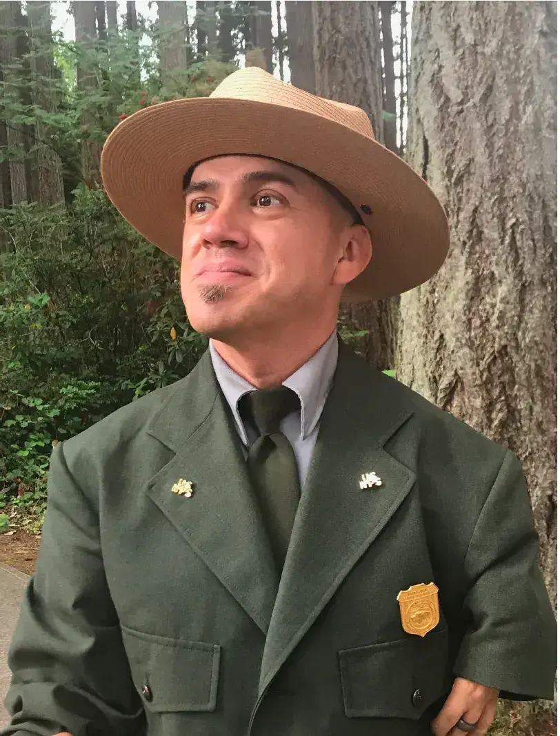 National Park Service ranger in uniform smiling near a tree in a forest setting.