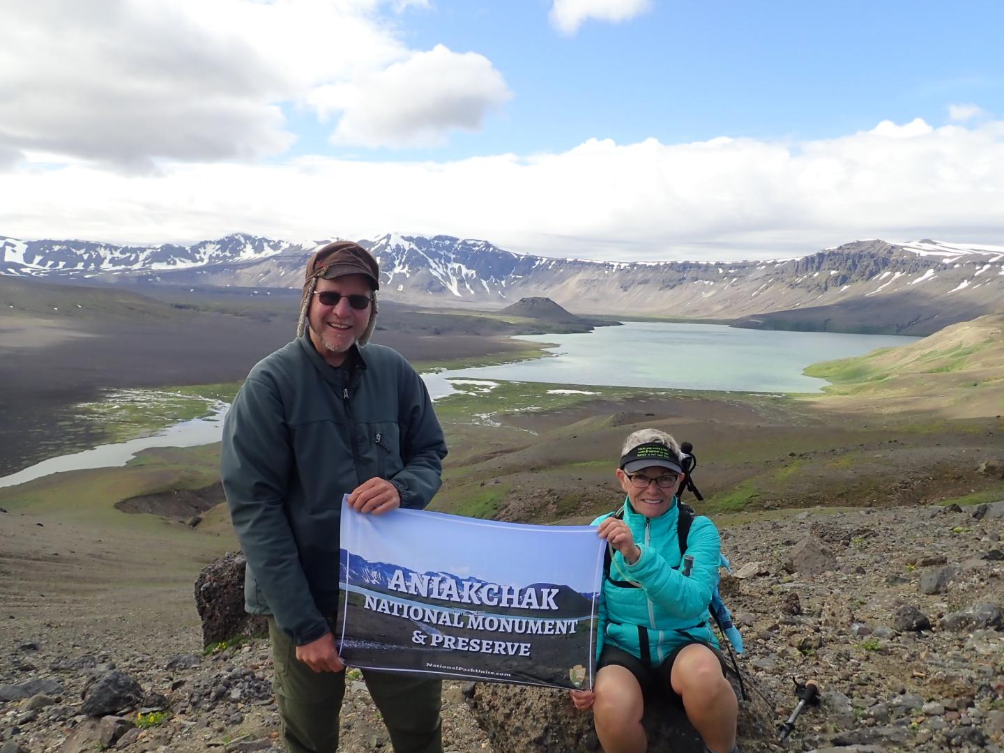Two white adults hold a banner on a rocky hill with a lake and mountains in the background.