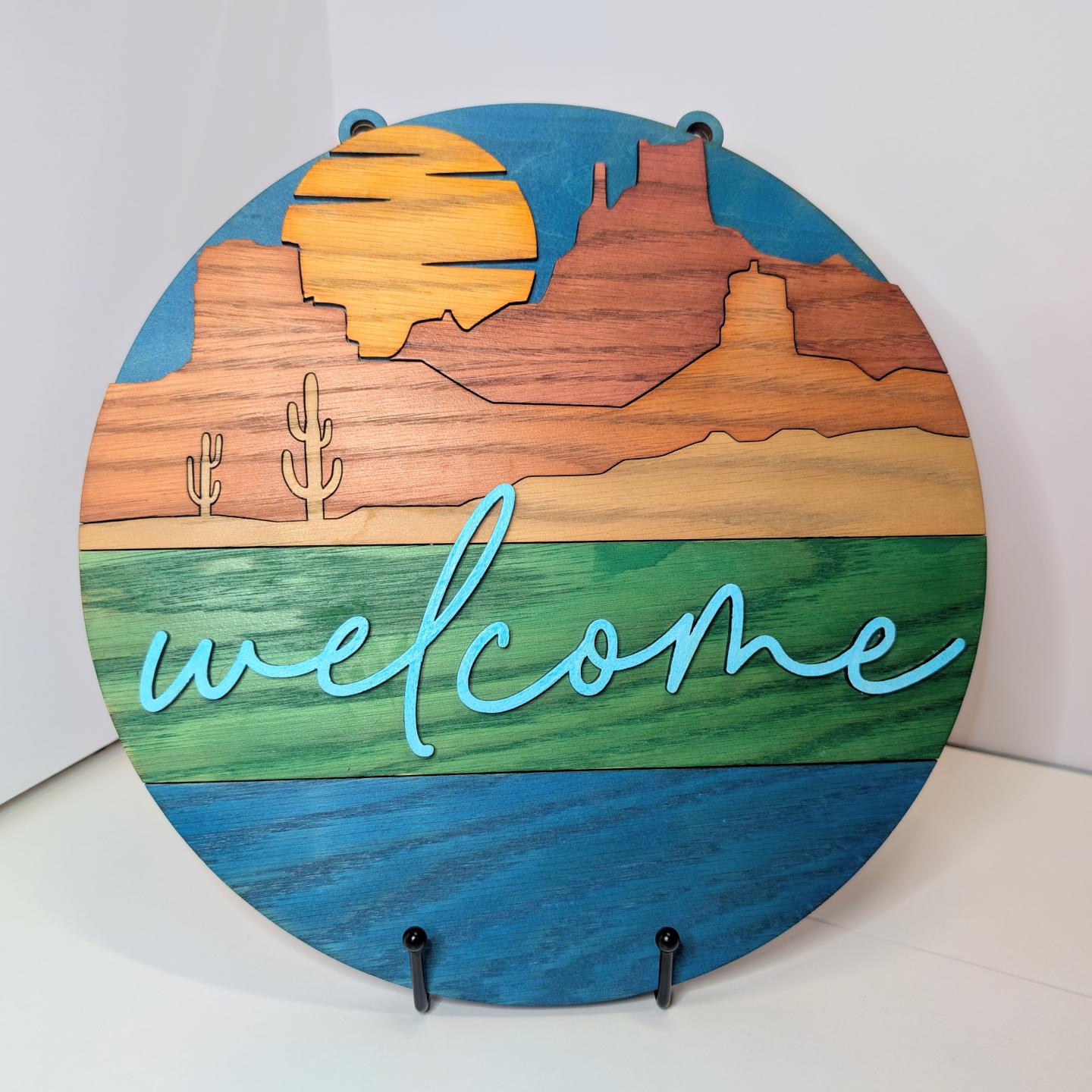 Round wooden plaque with desert scene, sun, cacti, and "welcome" in cursive.