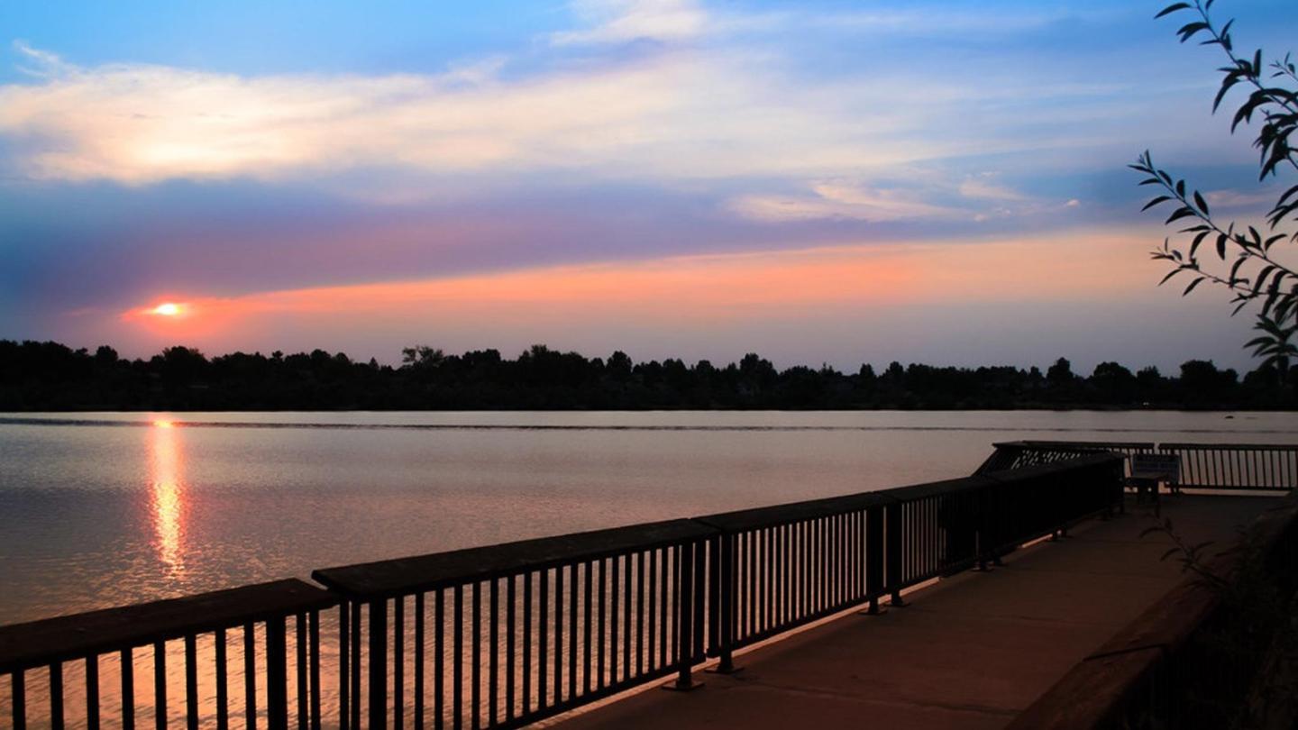 Sunset over a calm lake with a silhouetted railing and colorful sky.