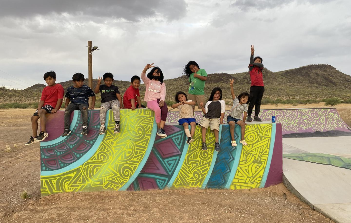 Children sitting on colorful graffiti-covered skate ramp, with mountains in the background.