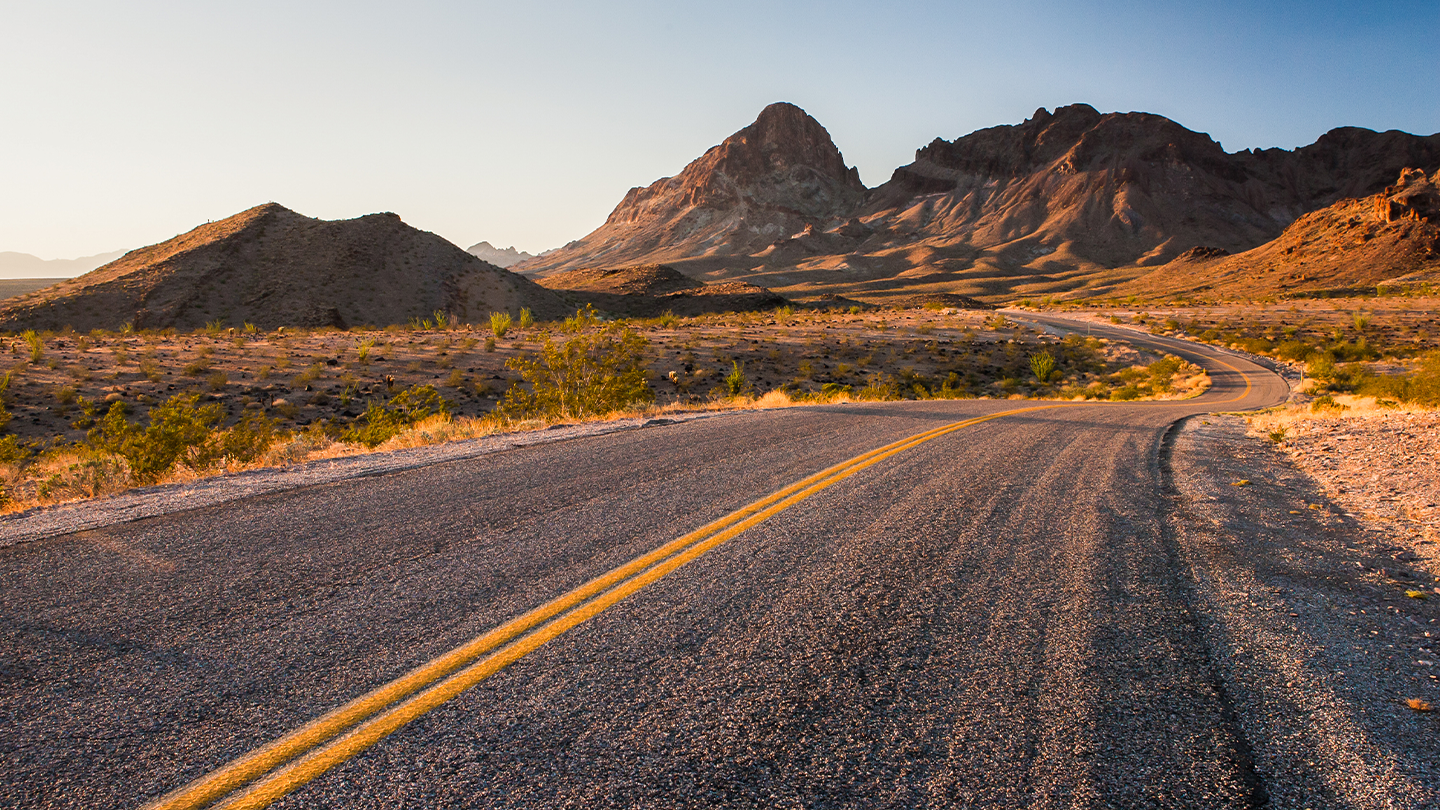 Curving desert road leading to distant mountains at sunset.