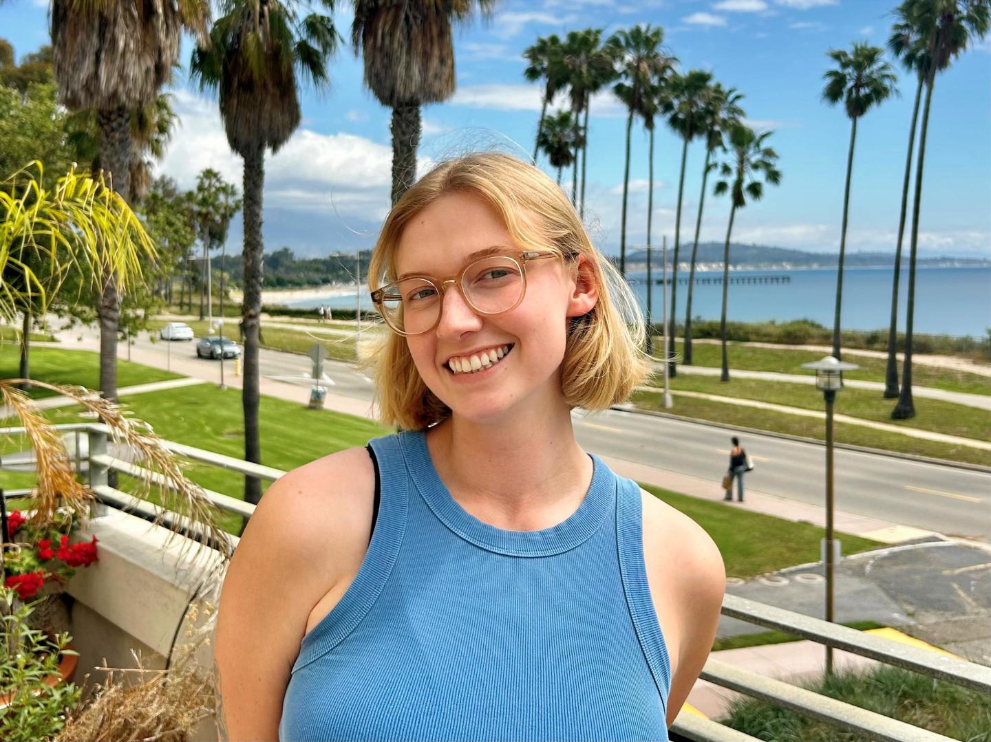 Smiling person with glasses, palm trees, ocean and blue sky in background.