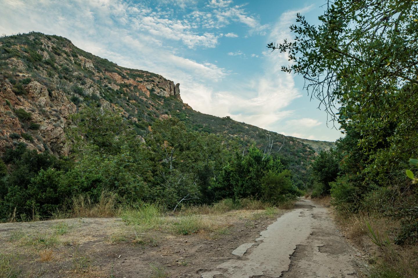 Dirt path through lush green valley under a cloudy blue sky.