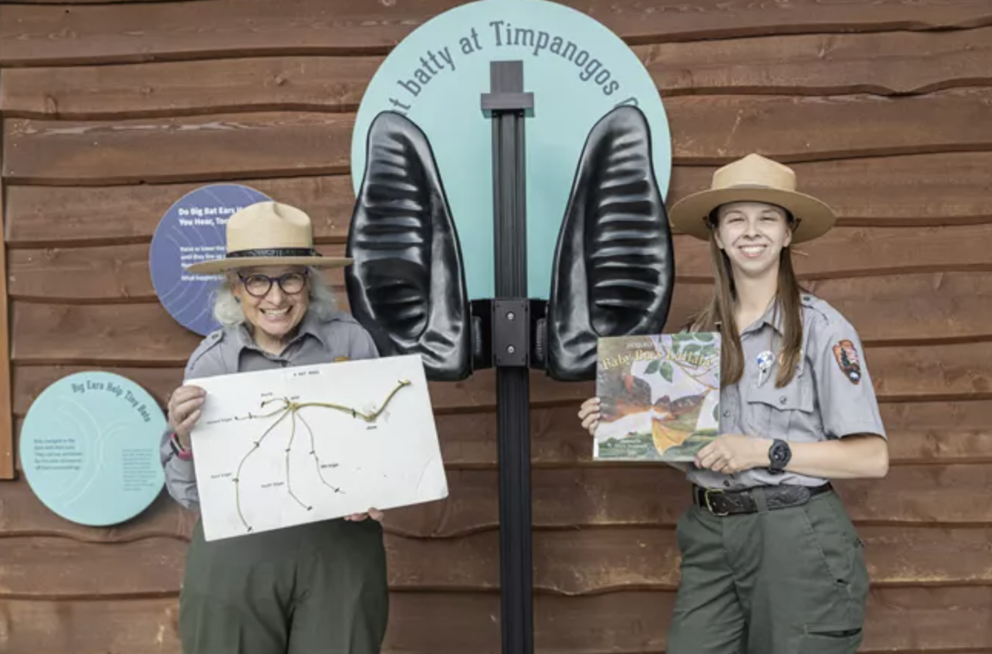 Two park rangers in uniform holding nature posters, standing beside a butterfly display.