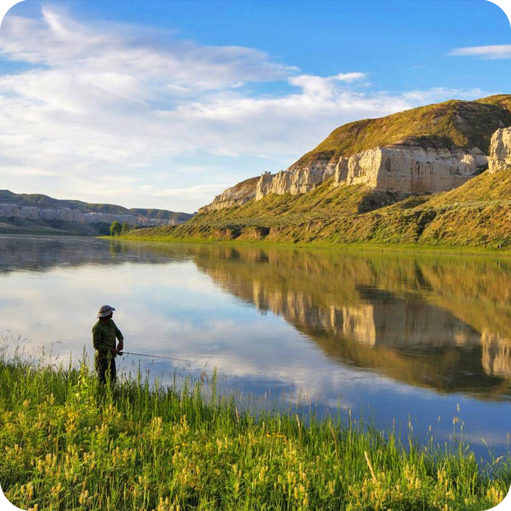 Person fishing by a calm river with cliffs in the background under a blue sky.