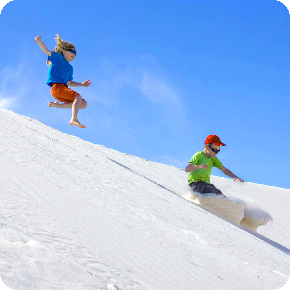 Two kids playfully running down a white sand dune under a bright blue sky.