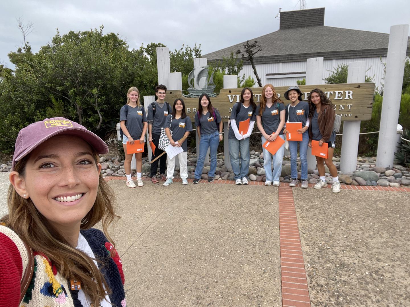 Group of smiling people outside a building, one taking a selfie, holding orange folders.