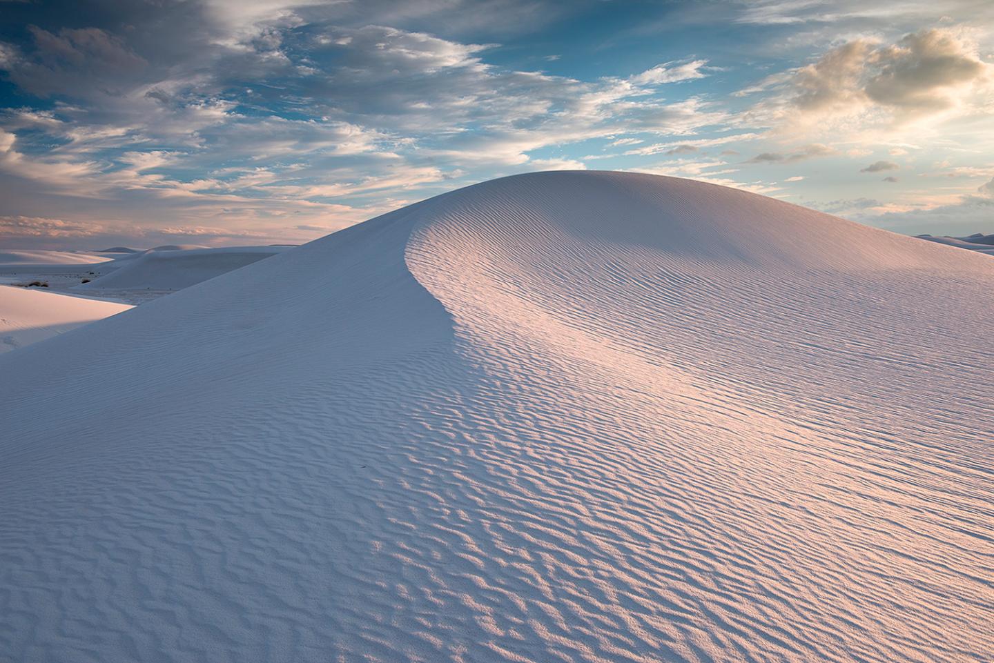 Rippling sand dune under a blue sky with clouds at sunset.