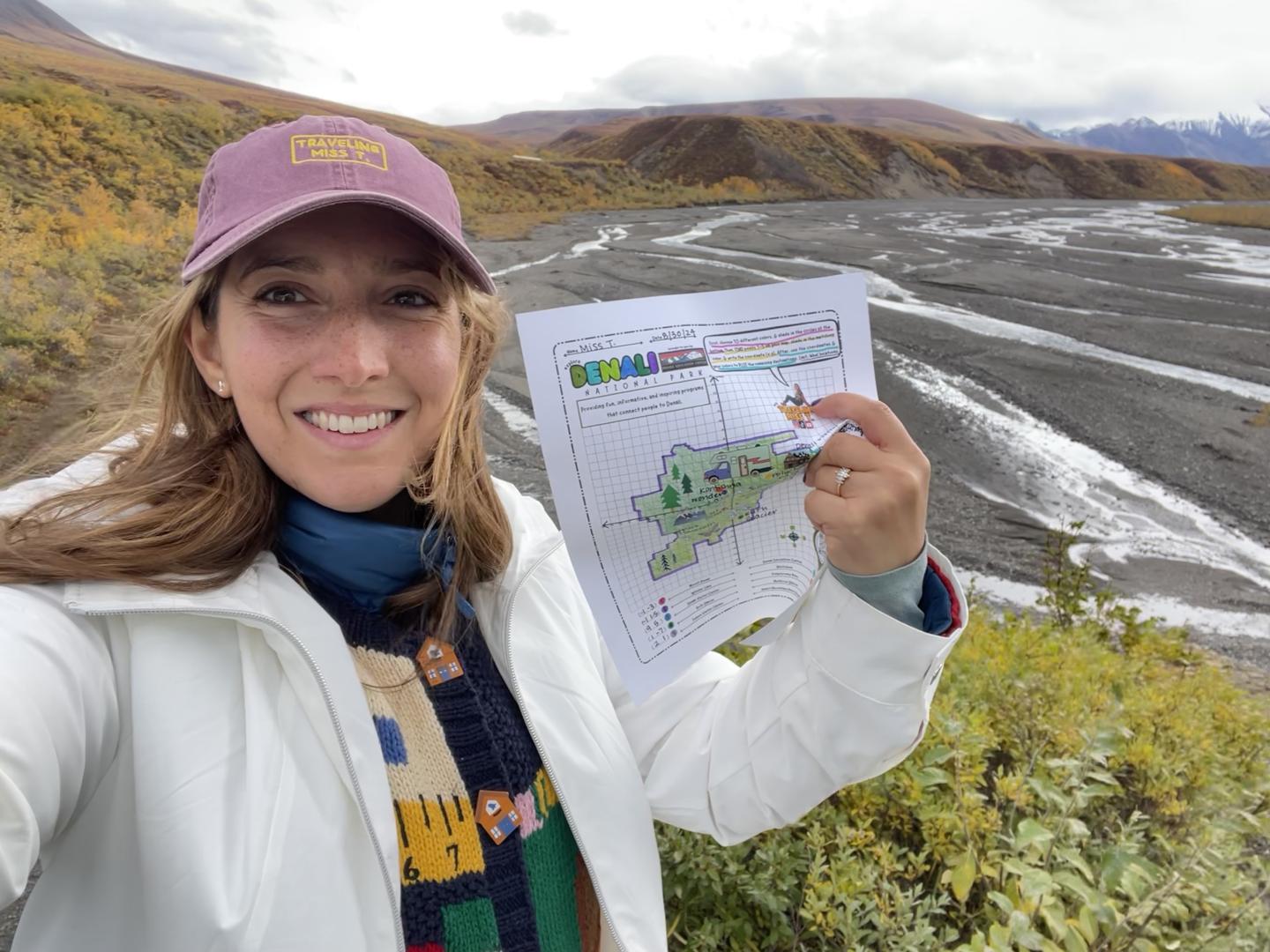 Smiling person holding a map in a scenic, mountainous landscape.