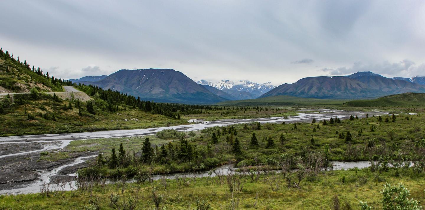 Mountain landscape with river, green fields, and cloudy sky.