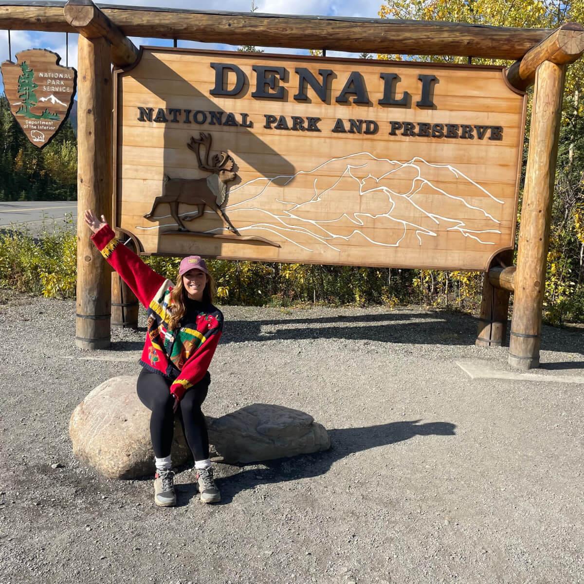 Person sitting by Denali National Park sign, pointing upwards.