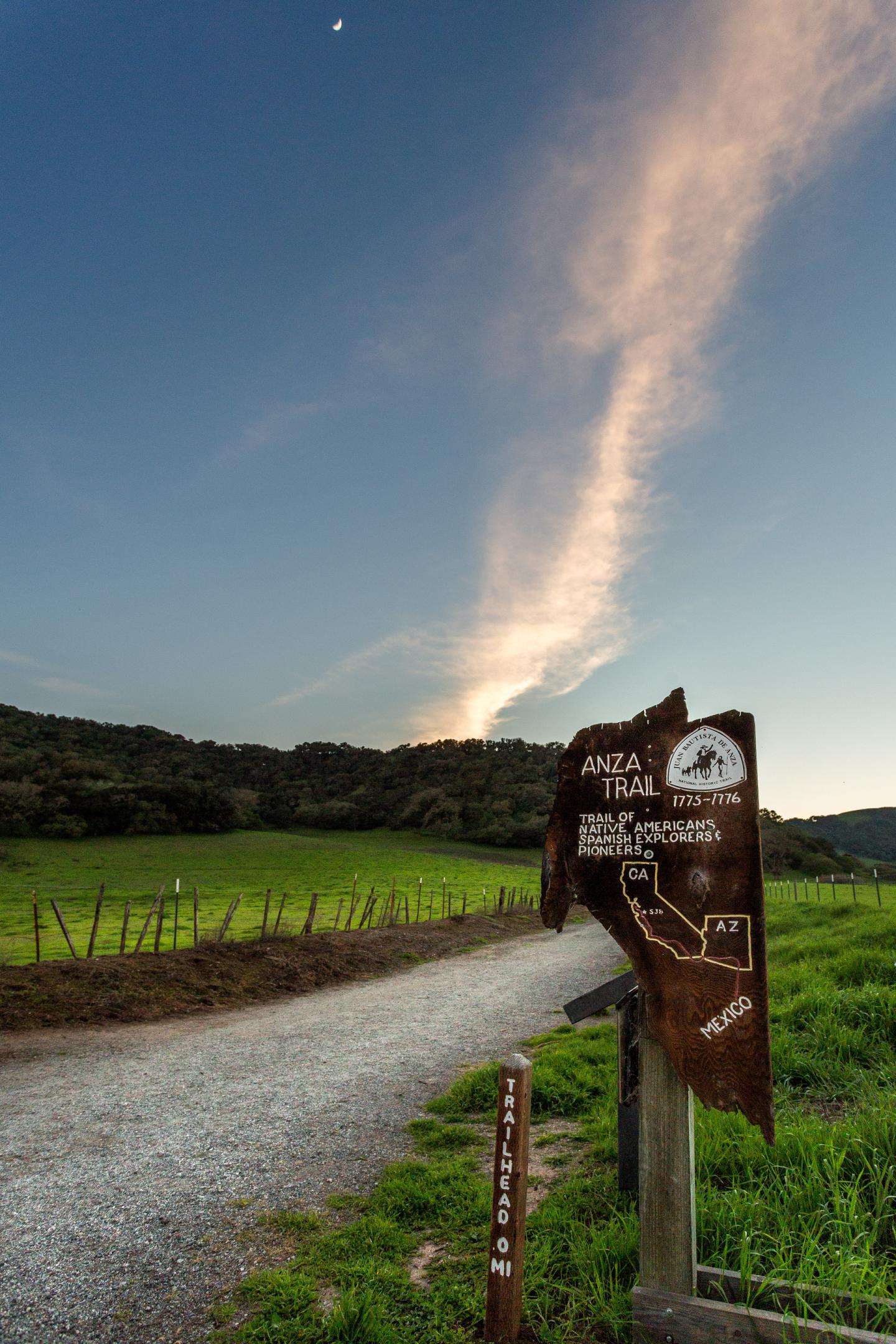 Trail sign on a dirt path at sunset with fields and hills in the background.