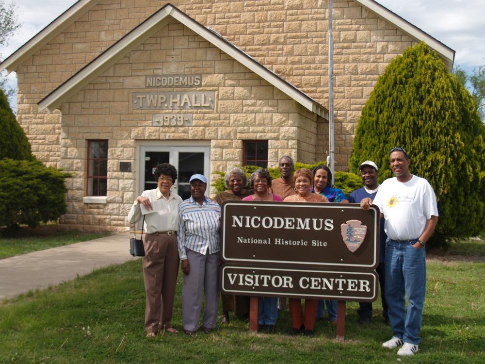 Group stands smiling by "Nicodemus Visitor Center" sign, stone building behind.
