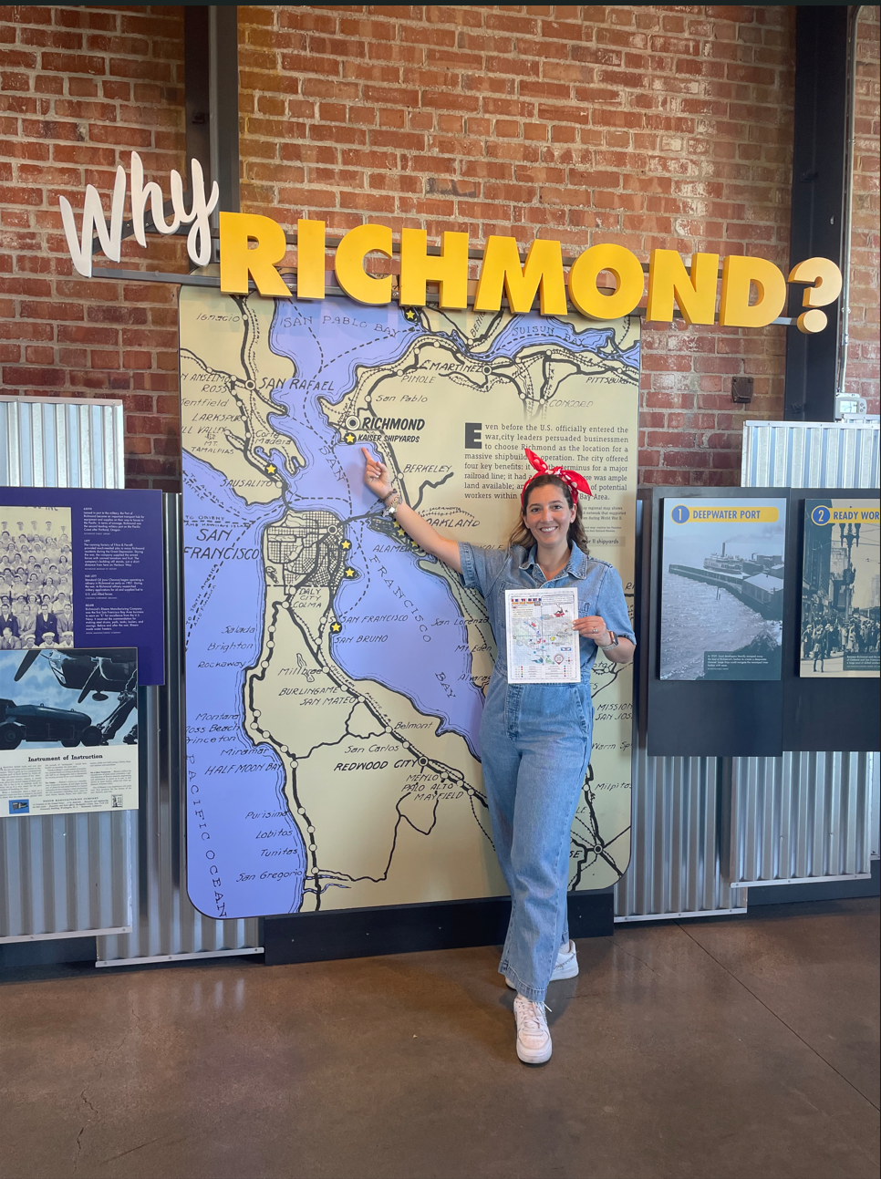 Person in denim outfit and red bandana points at a large map display indoors.