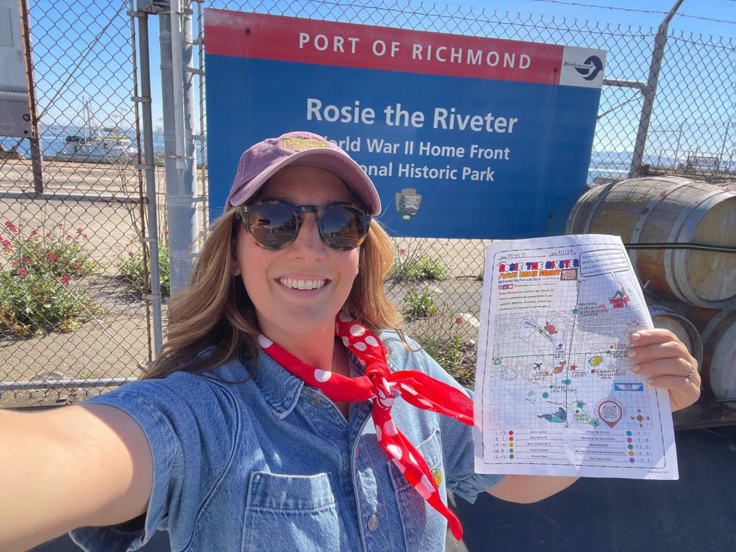 Woman smiling in front of a "Rosie the Riveter" sign, holding a map, wearing a cap and scarf.