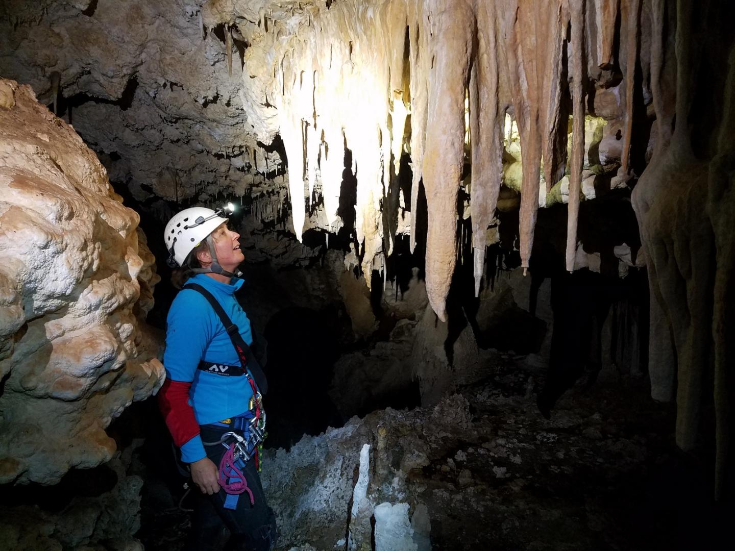 Caver in blue jacket, helmet, among stalactites in a dimly lit cave.