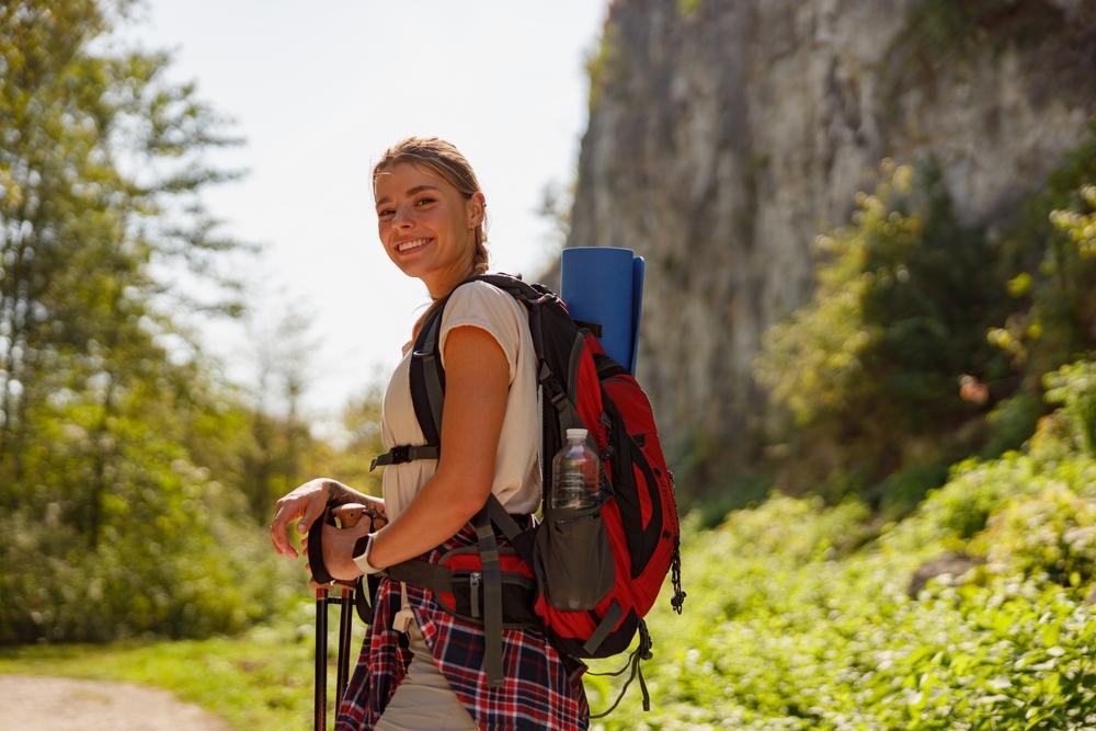 Smiling hiker with a backpack on a sunny trail near a rocky cliff.