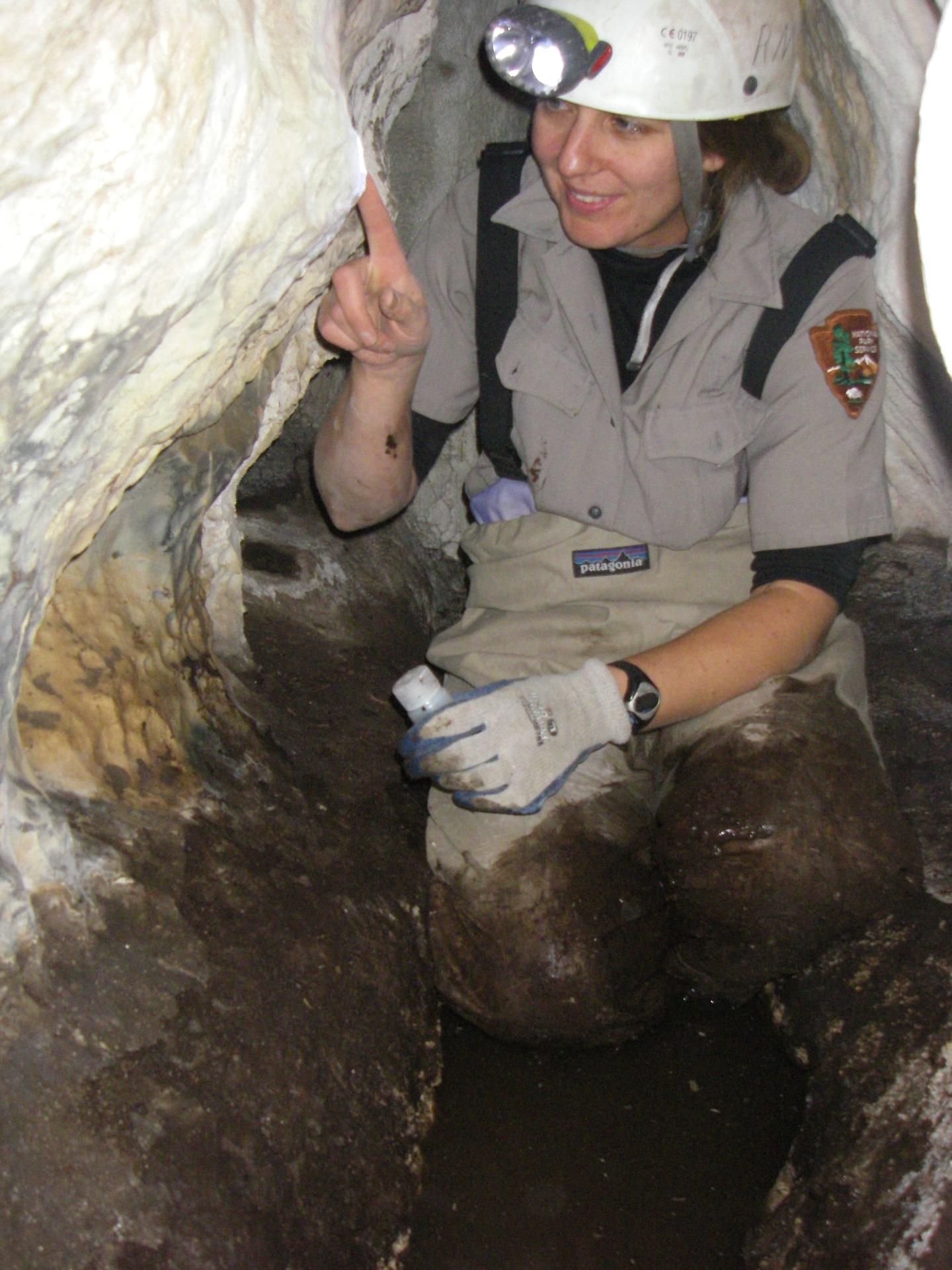 Explorer in cave kneeling, wearing a helmet and gloves, pointing at cave wall.