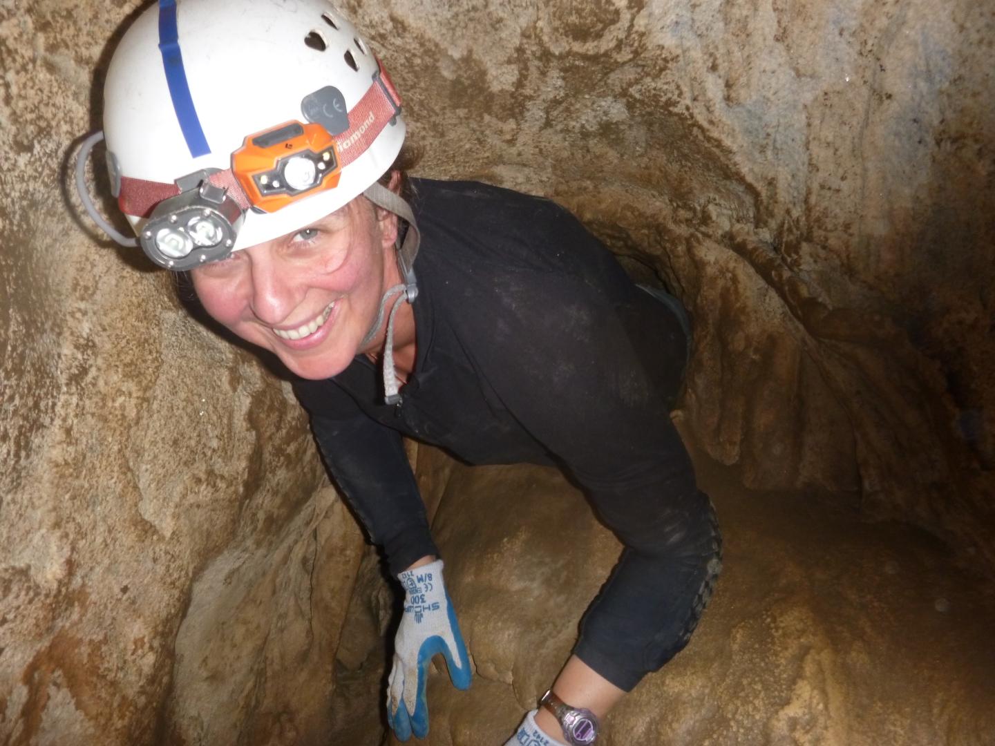 Caver wearing helmet and gloves, smiling in a narrow cave passage.