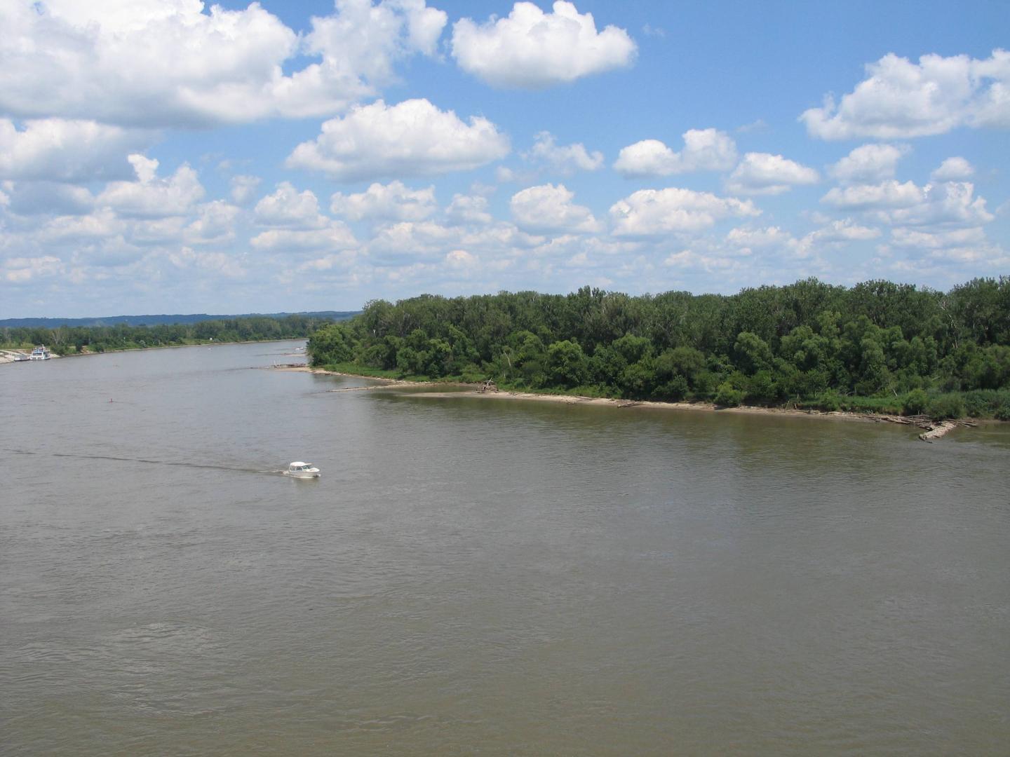 River with a small boat, trees, and a cloudy blue sky.
