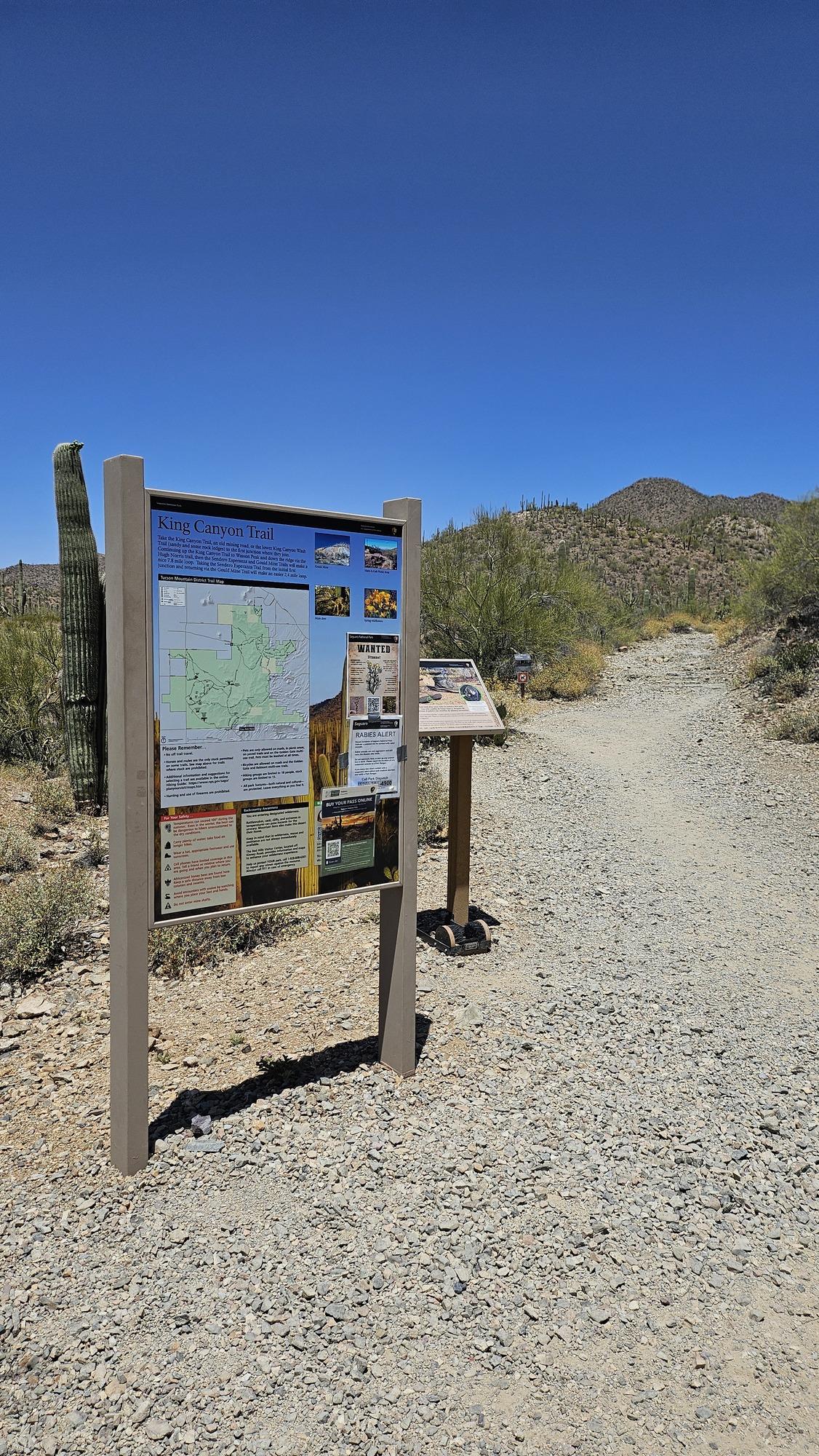Trailhead sign in a desert landscape under clear blue sky.