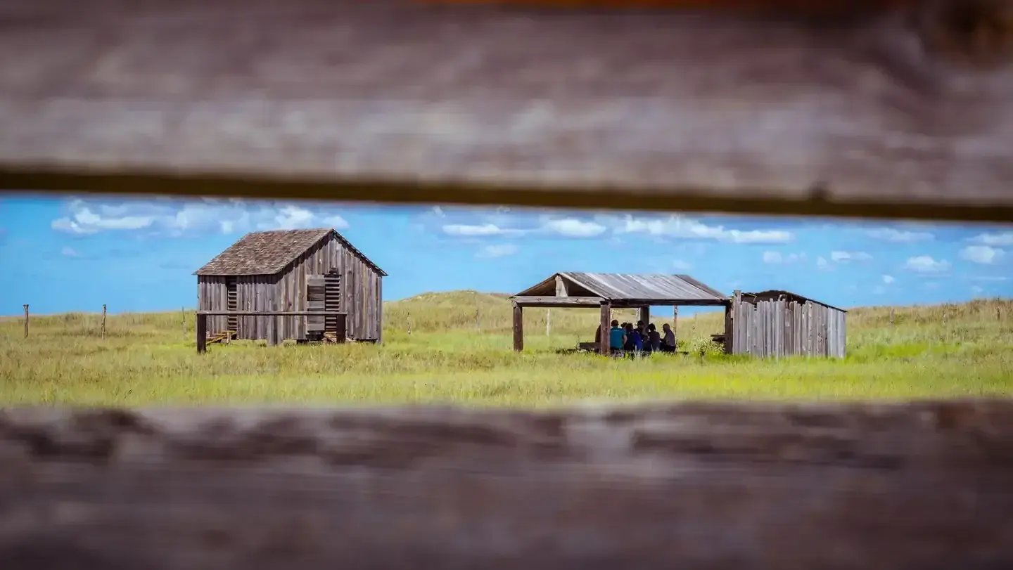 Novillo Line Camp gives view of ranching history of Padre Island National Seashore on grassy field under blue sky, seen through a fence gap.