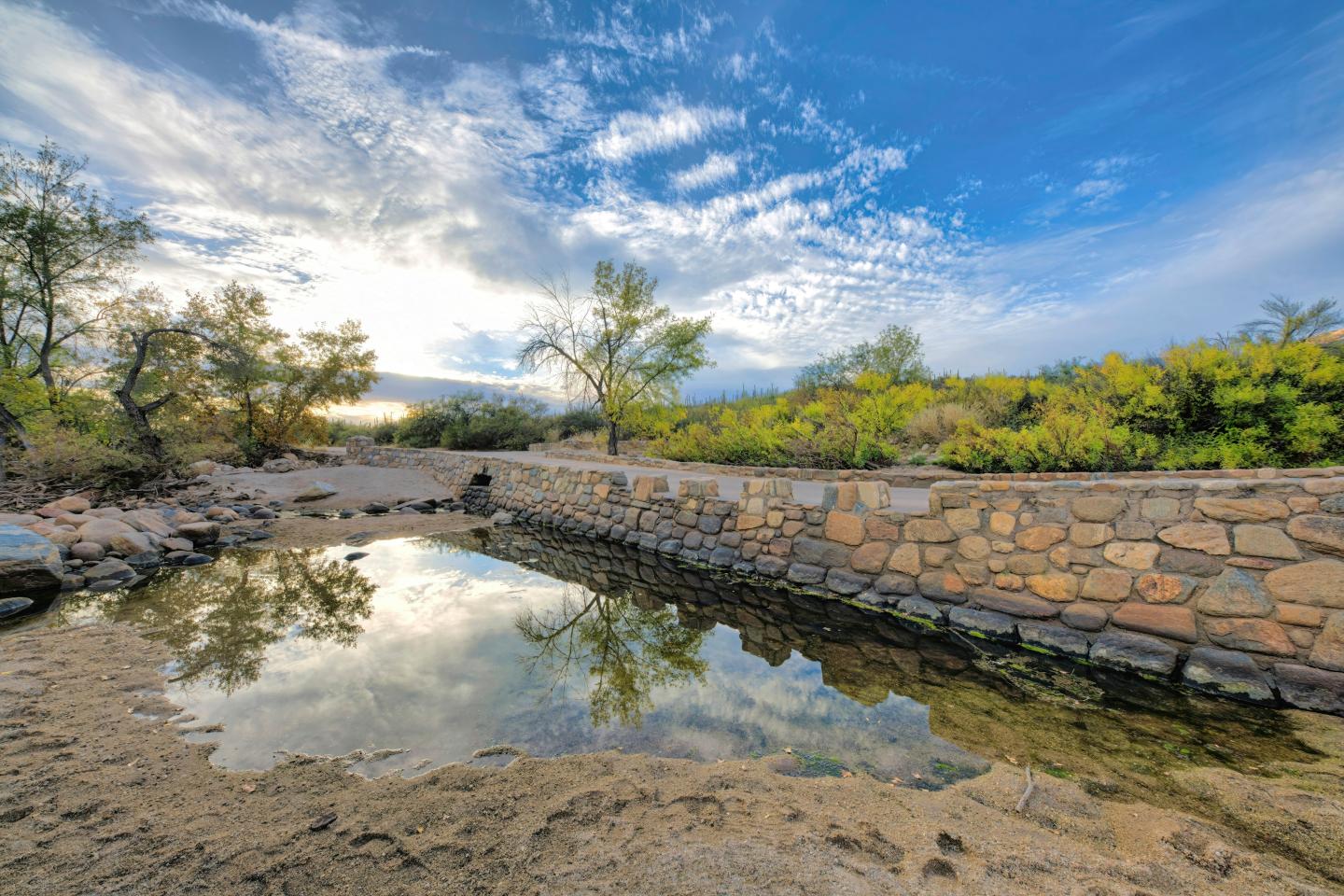 Stone bridge over a calm river with a cloudy sky and green foliage.