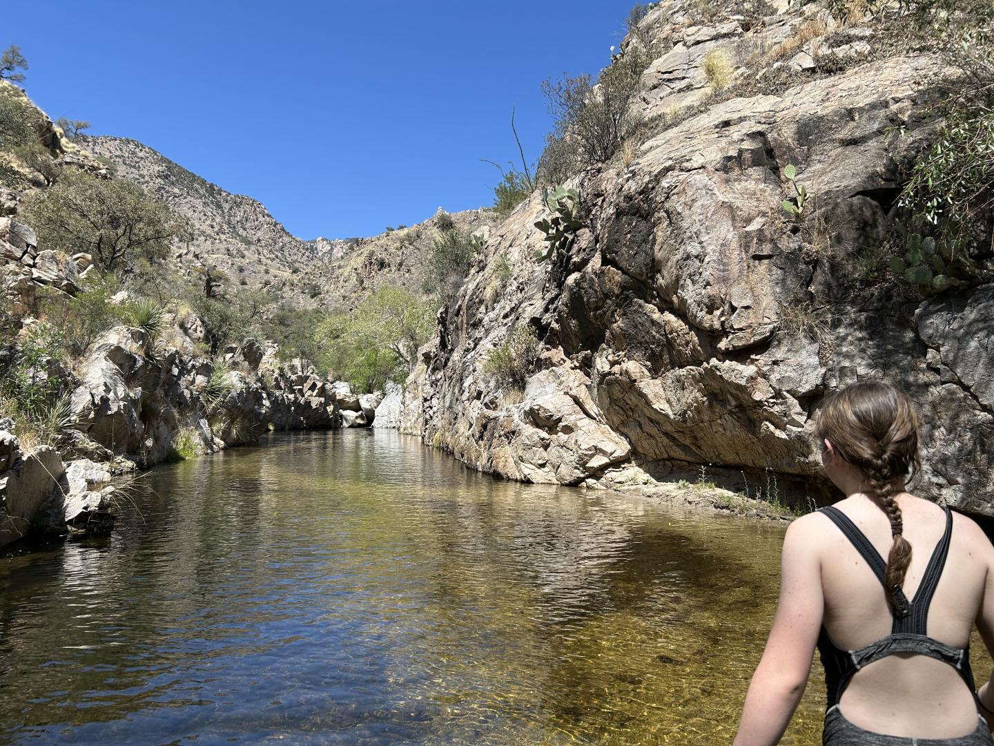 Person standing by a clear stream, surrounded by rocky cliffs under a blue sky.